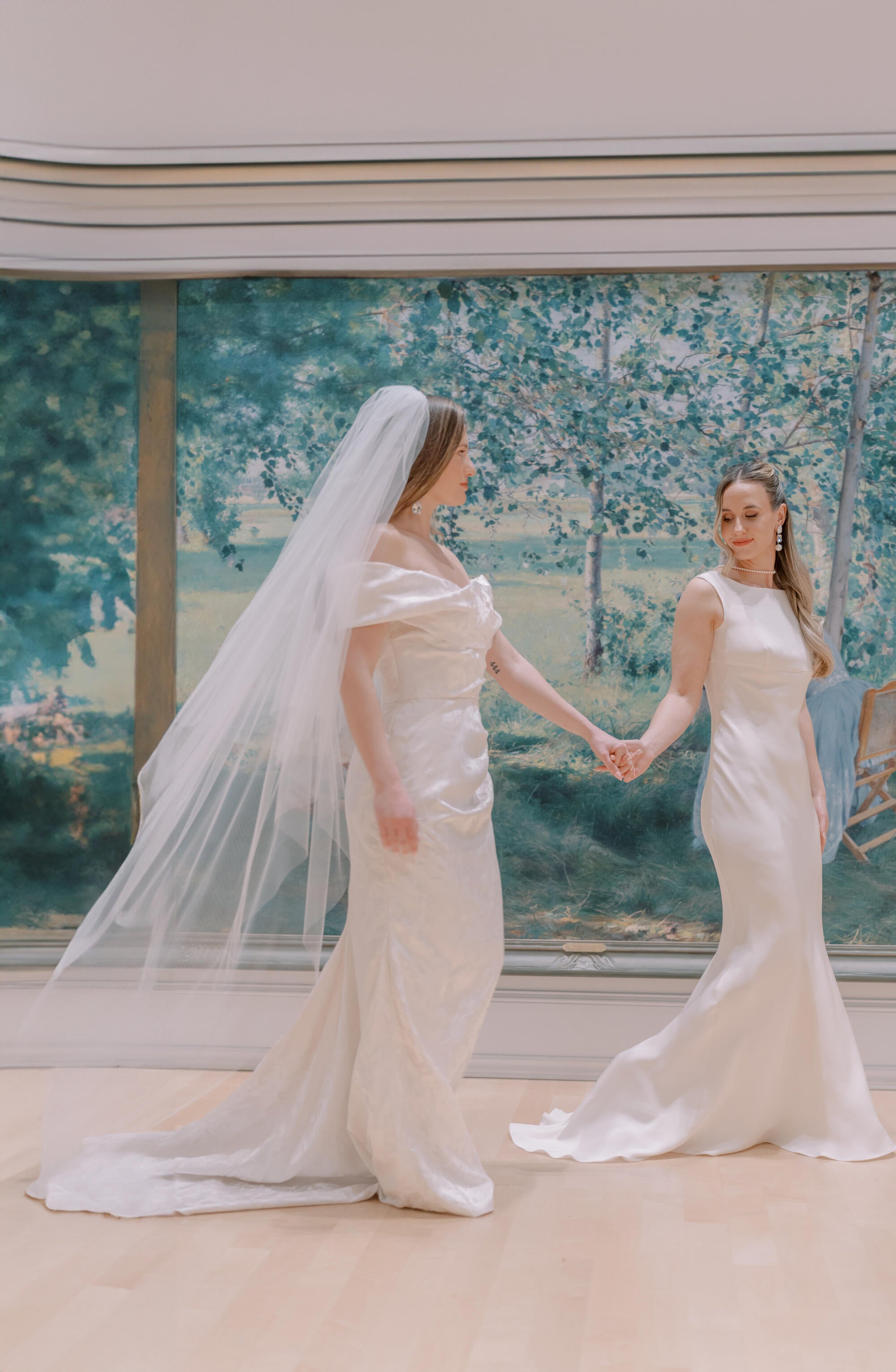 Two women in wedding dresses holding hands in front of a decorative wall.