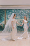 Two women in wedding dresses holding hands in front of a decorative wall.