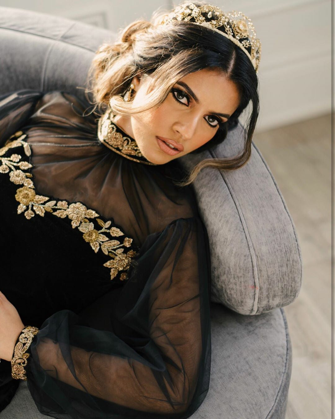 Woman wearing a black outfit with gold embroidery and a headpiece, sitting on a gray couch.