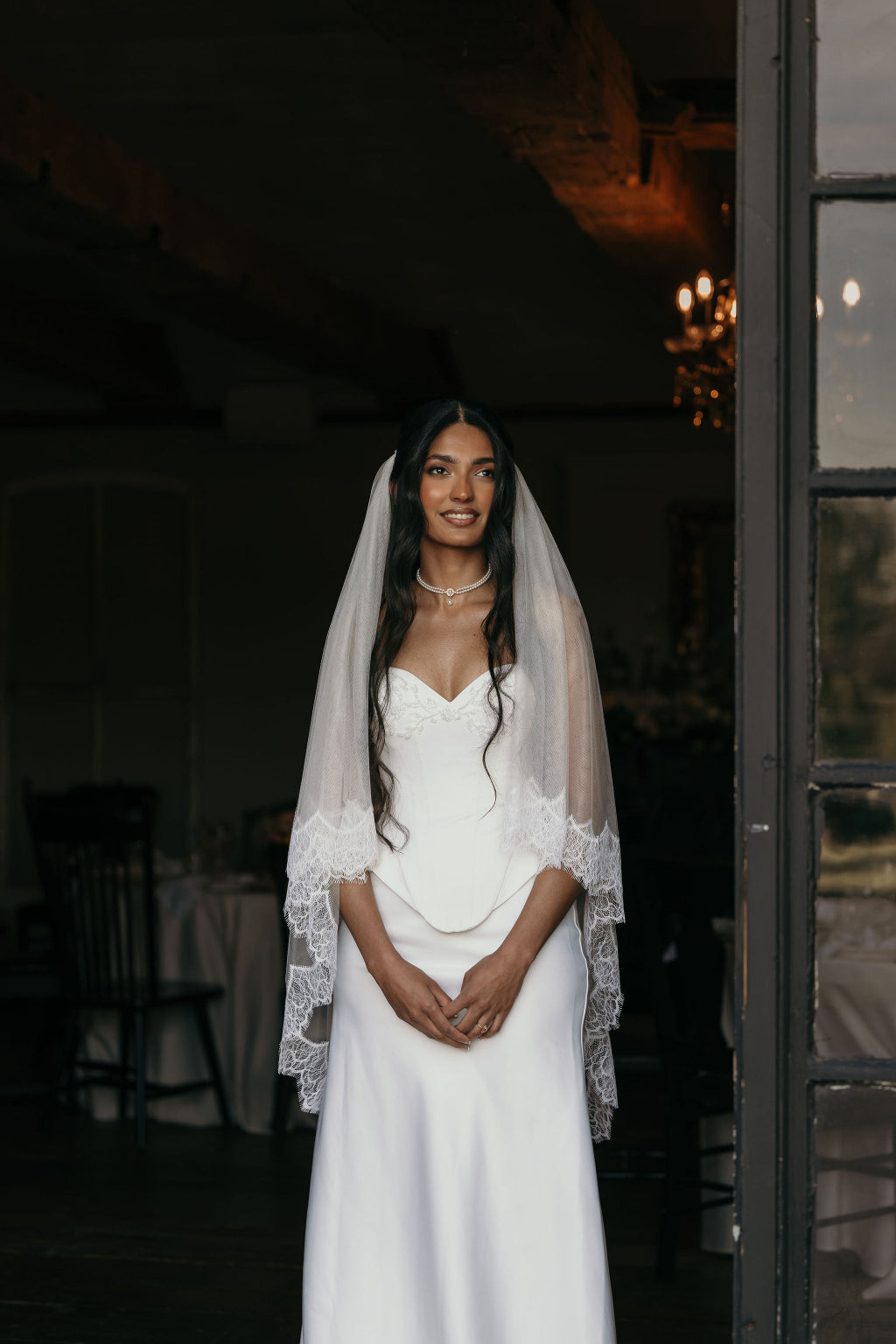 Woman in a white wedding dress with a veil standing in a dimly lit room.