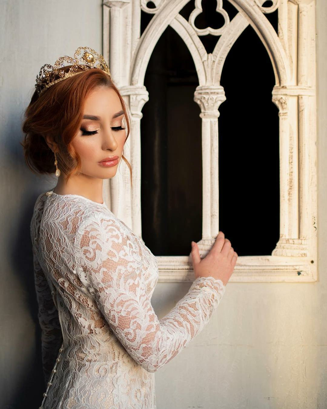 Woman in a lace dress and tiara standing in front of an ornate window.