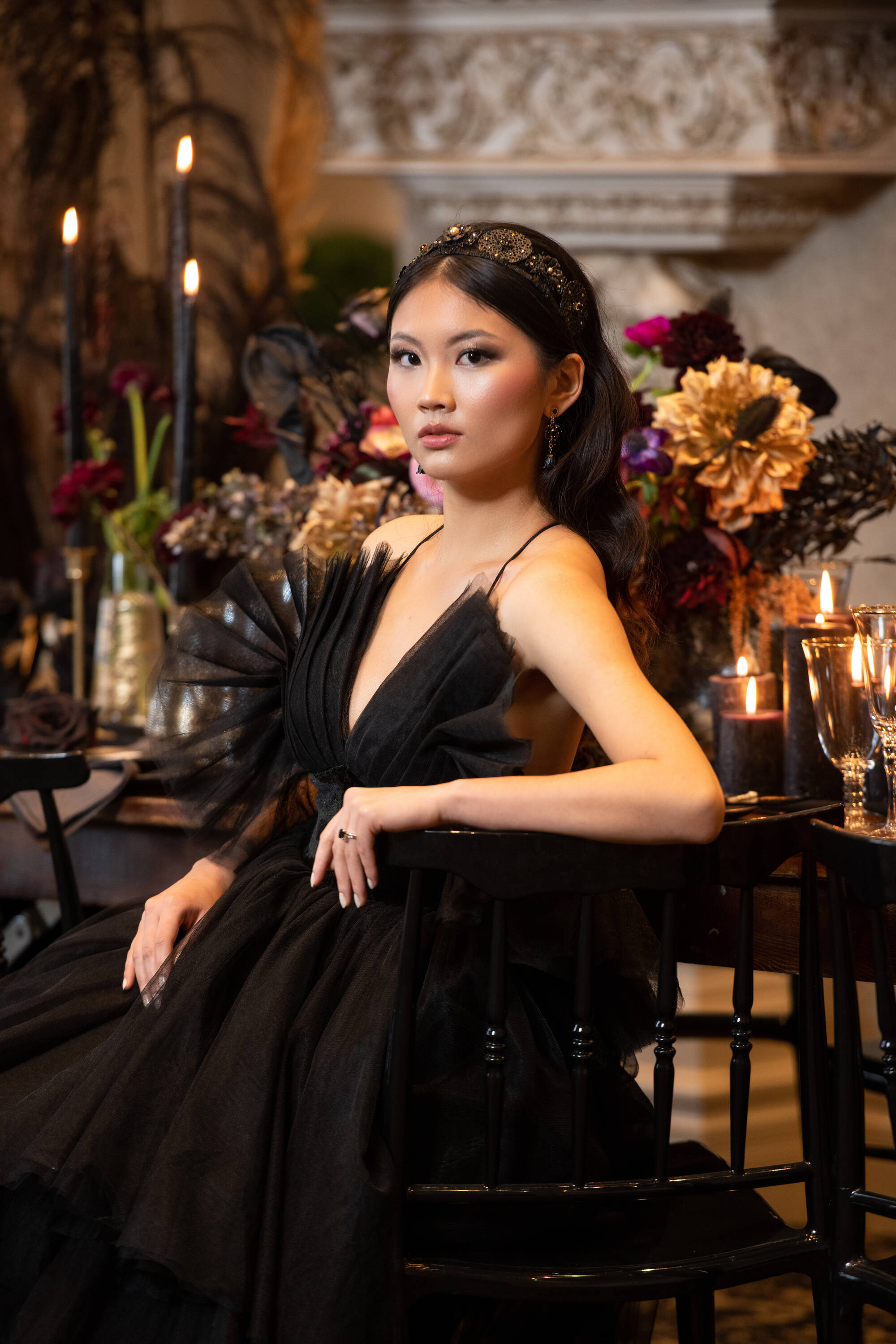 Woman in a black dress sitting at a table with floral arrangements and candles in an elegant setting.