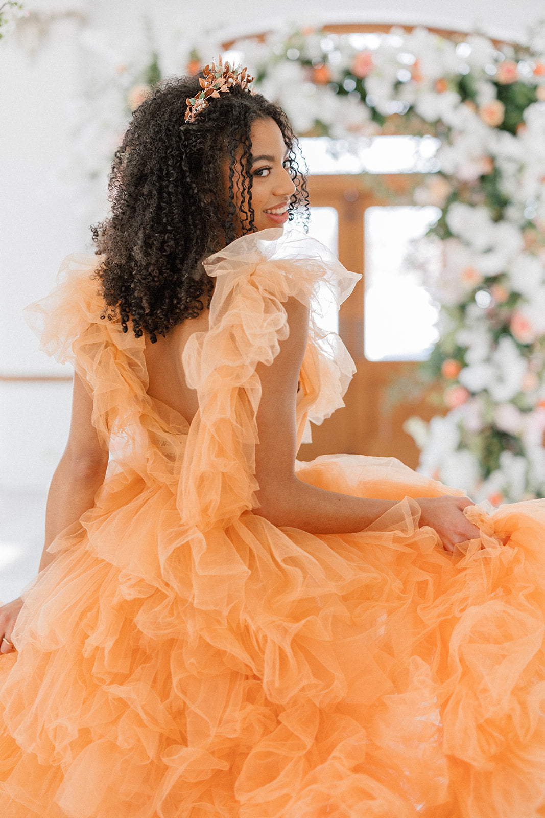 Woman in an orange dress with ruffled sleeves and a floral headpiece, sitting in a decorated indoor setting.
