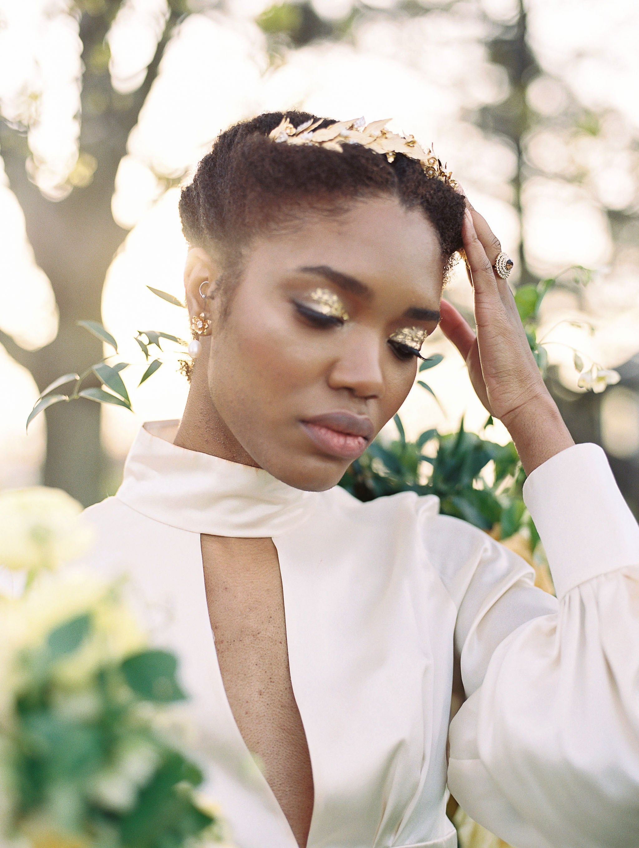 Woman in a white outfit with gold makeup and hair accessories, standing outdoors with greenery in the background.
