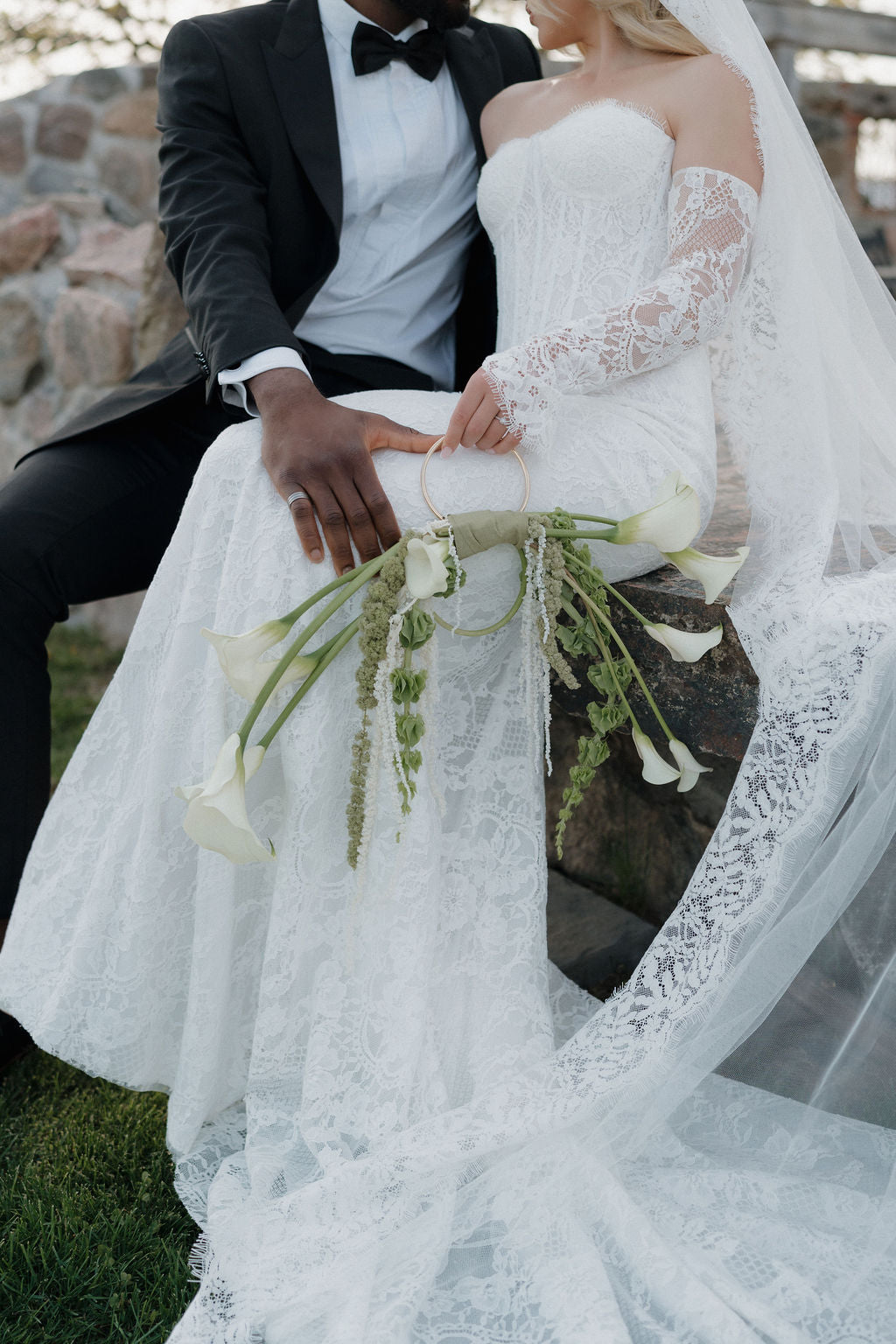 Couple in wedding attire sitting outdoors with a stone wall background