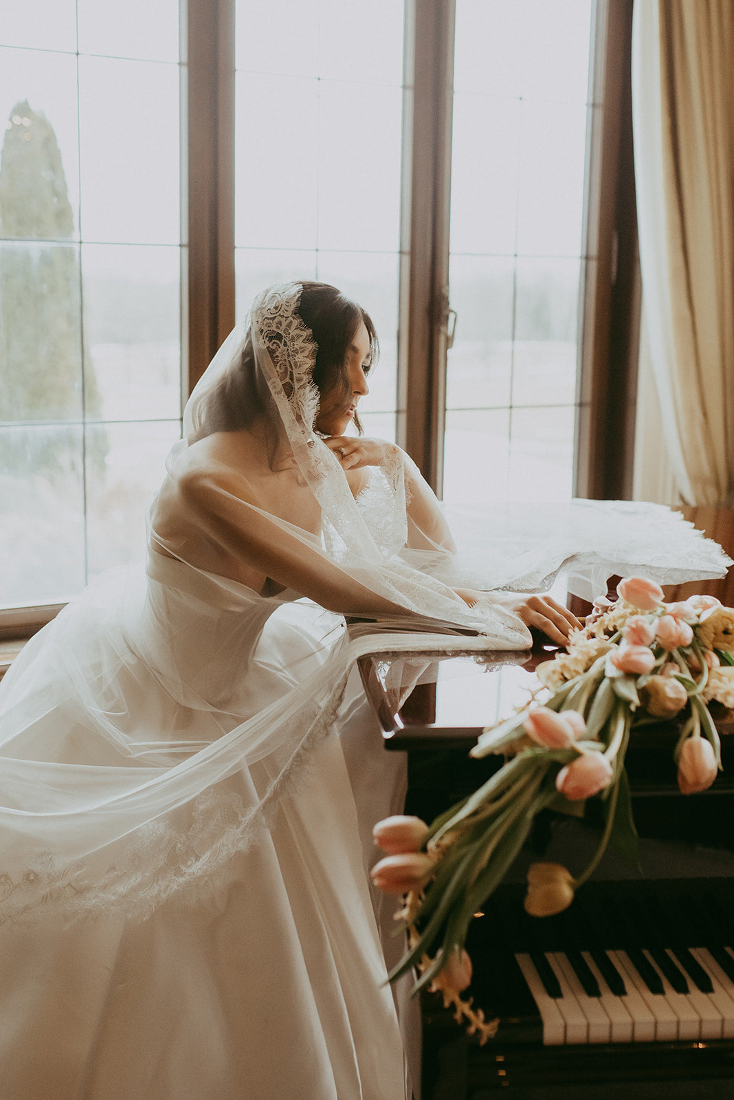 Bride in a white gown with a veil sitting at a piano with flowers in front of her.