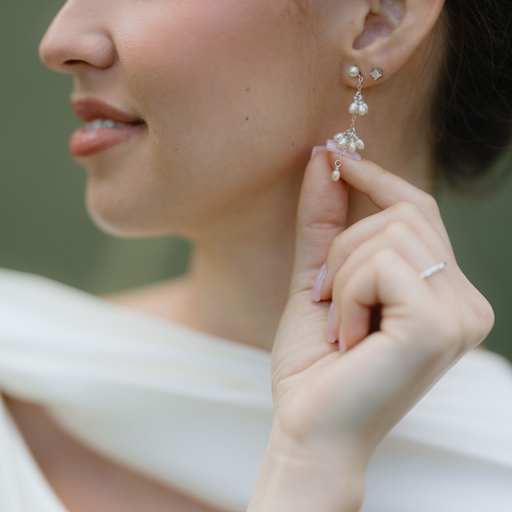 A close-up lifestyle shot of a woman wearing delicate silver drop earrings with clear crystal-like beads and a matching silver ring, with a white dress in the background.