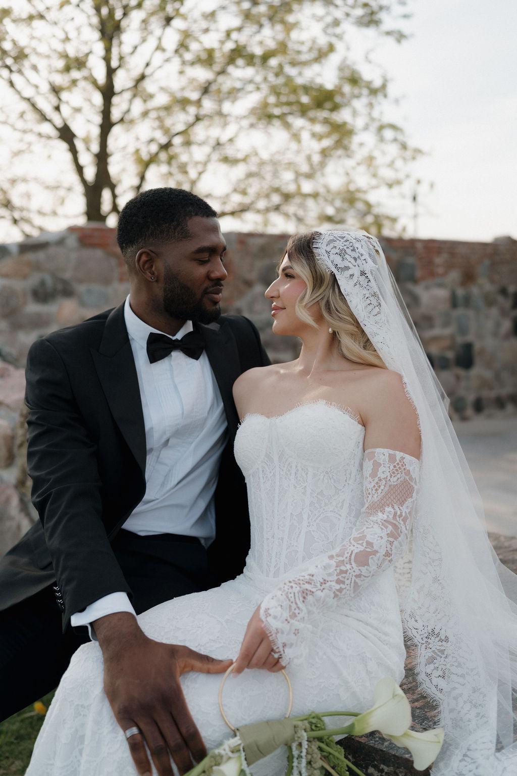 Couple in wedding attire sitting together with a stone wall and trees in the background