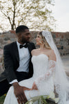Couple in wedding attire sitting together with a stone wall and trees in the background