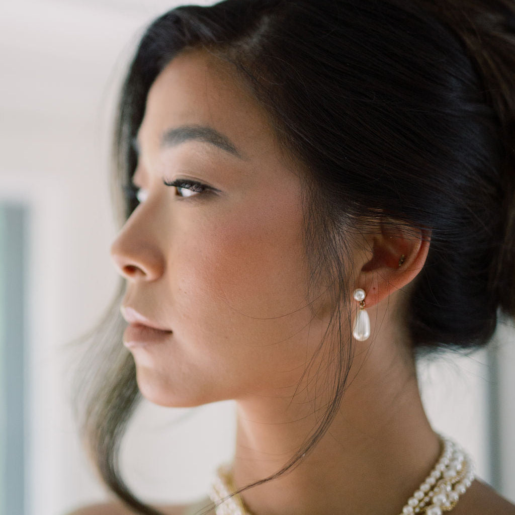 A close-up side profile of a woman with dark hair in a bun, wearing a multi-strand pearl necklace and a matching pearl drop earring.