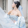Woman in a light blue gown with floral details standing in front of classical architecture.