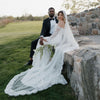 Couple in wedding attire sitting on a stone wall outdoors.