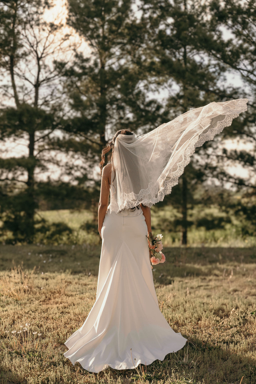 Woman in a white wedding dress with a long veil standing in a field with trees in the background