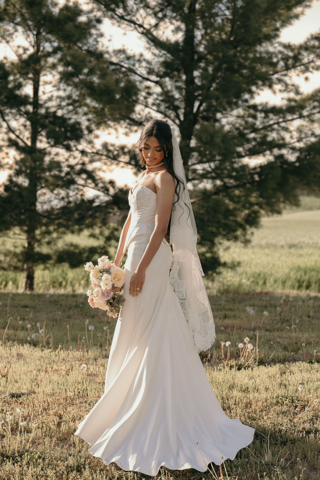 Woman in a white wedding dress standing in a field with trees in the background
