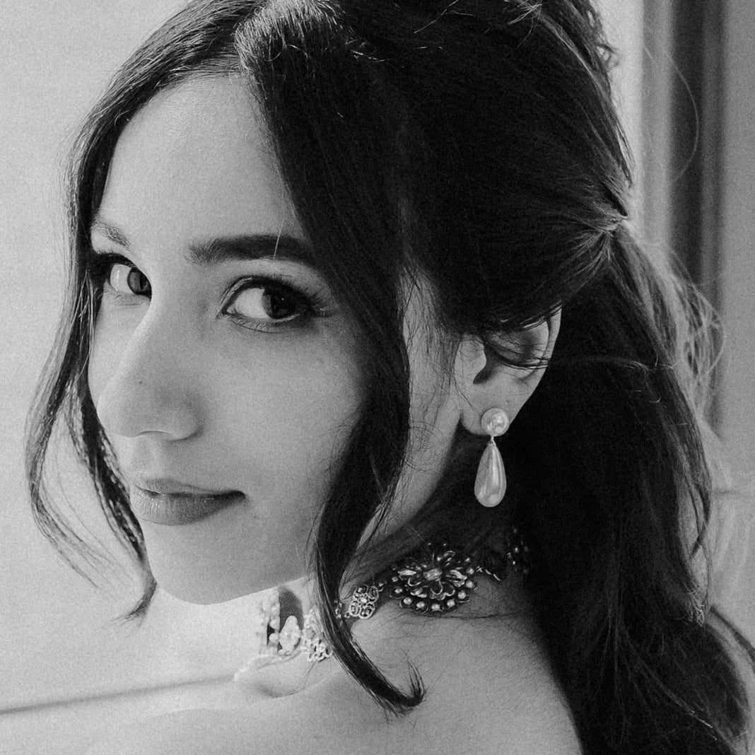 Black and white portrait of a woman with dark hair, wearing a pearl drop earring and a beaded choker, looking back over her shoulder.