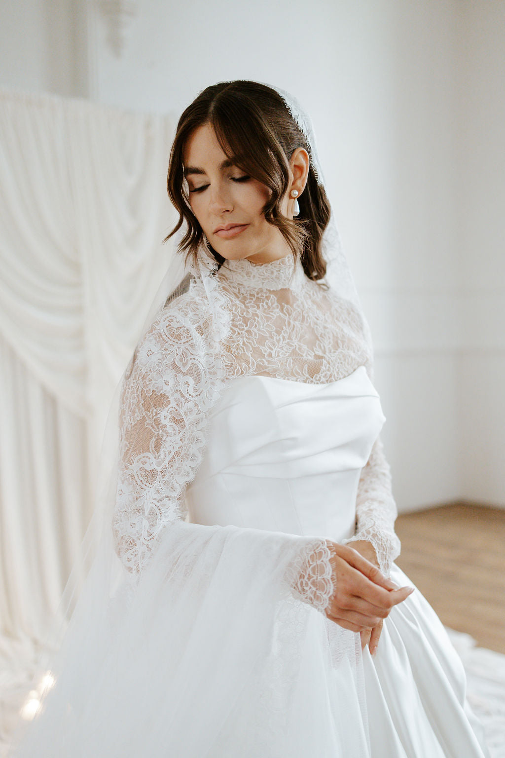 A woman wearing a white bridal gown with a sheer lace long-sleeve overlay and a full tulle skirt, posed indoors against a soft white draped backdrop.