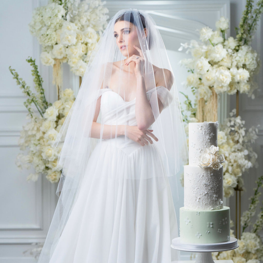 Bride in a white wedding dress with a long veil standing next to a wedding cake in a decorated room.