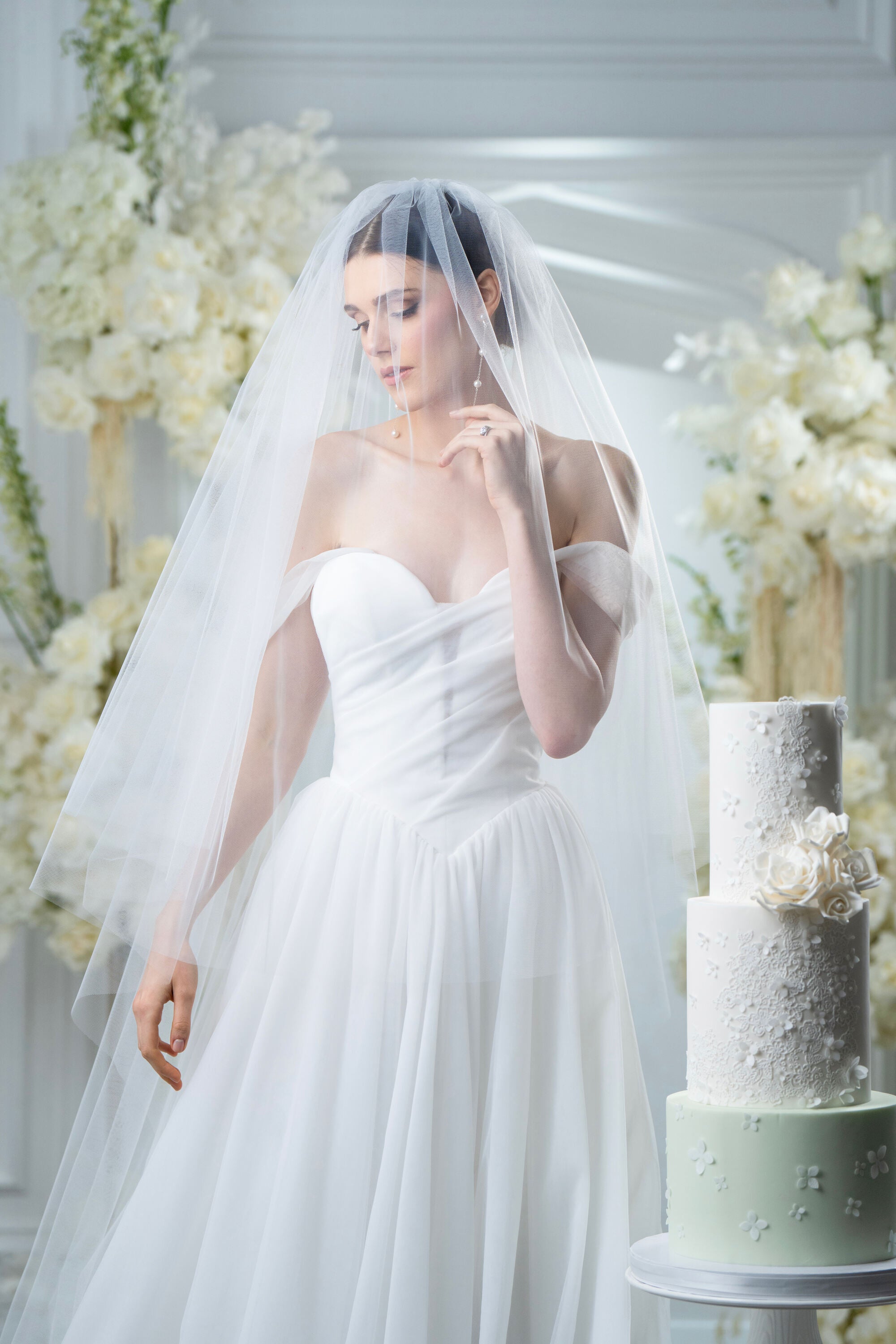 Woman in a white wedding dress with a veil, standing next to a wedding cake in a decorated room.