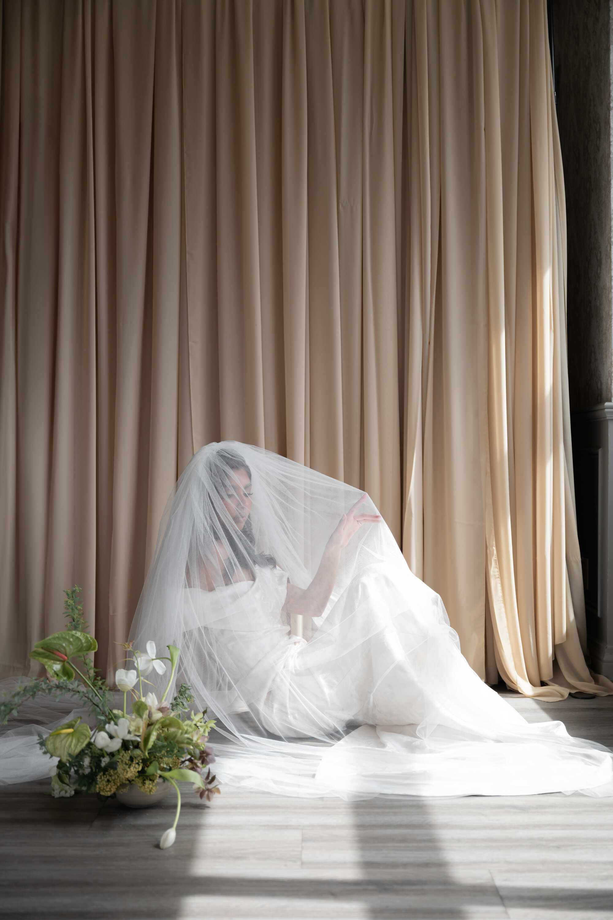 Bride in a white wedding dress sitting on the floor with a veil, surrounded by flowers against a beige curtain.