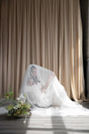 Bride in a white wedding dress sitting on the floor with a veil, surrounded by flowers against a beige curtain.