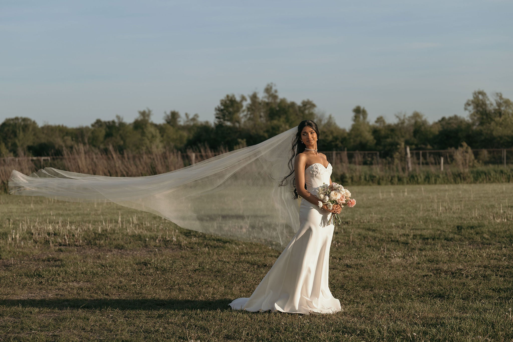 A bride in a strapless white wedding gown stands in an open grassy field with her long veil blowing in the wind, holding a bouquet of flowers. Trees and a fence are visible in the background under a pale blue sky.