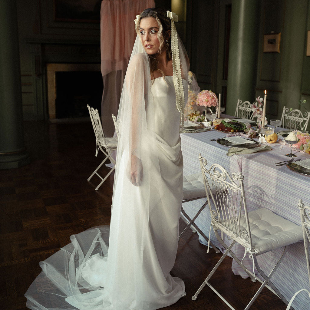 A bride in a strapless white wedding gown with a long sheer veil stands in a dimly lit indoor venue beside a decorated dining table with white chairs and pink floral arrangements.