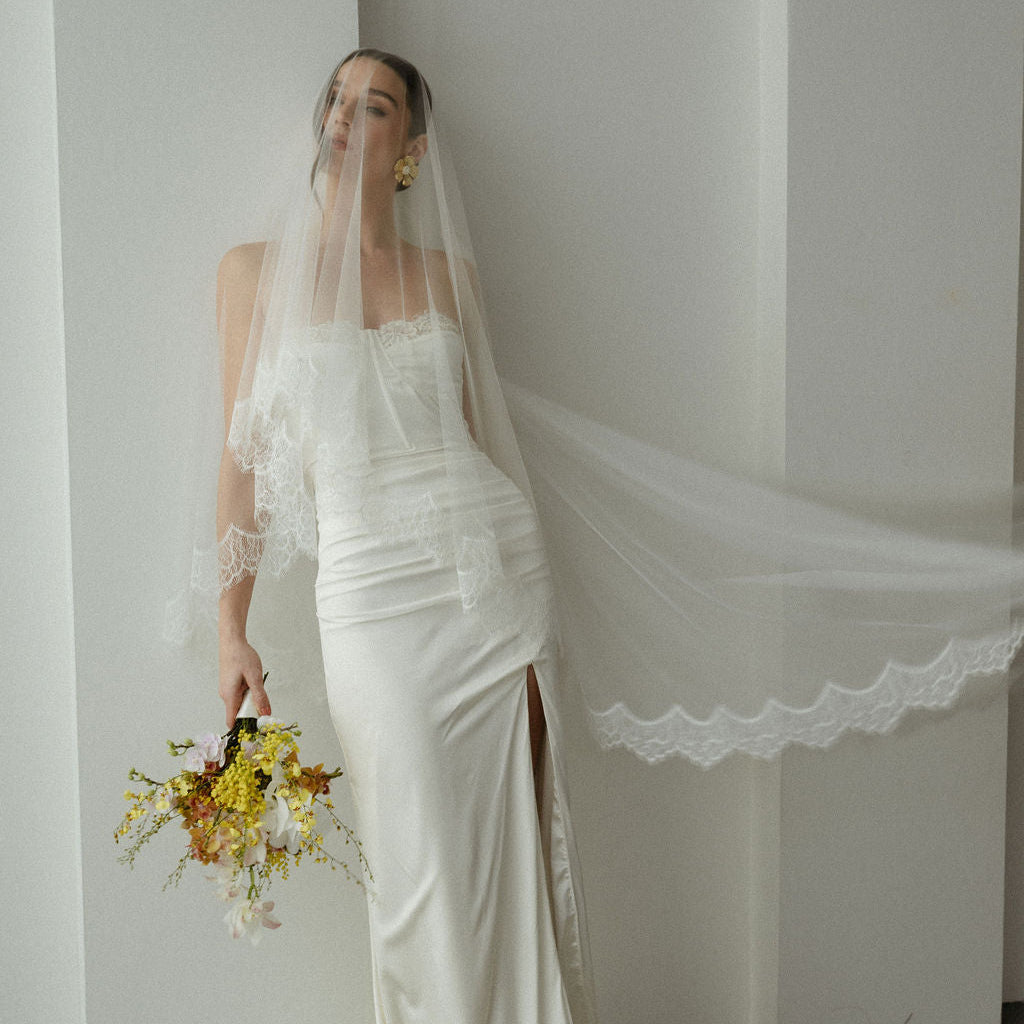 Woman in a white wedding dress with a long veil holding a bouquet against a plain background