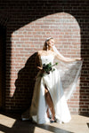Bride in a strapless wedding dress with a high slit, holding a bouquet against a brick wall.
