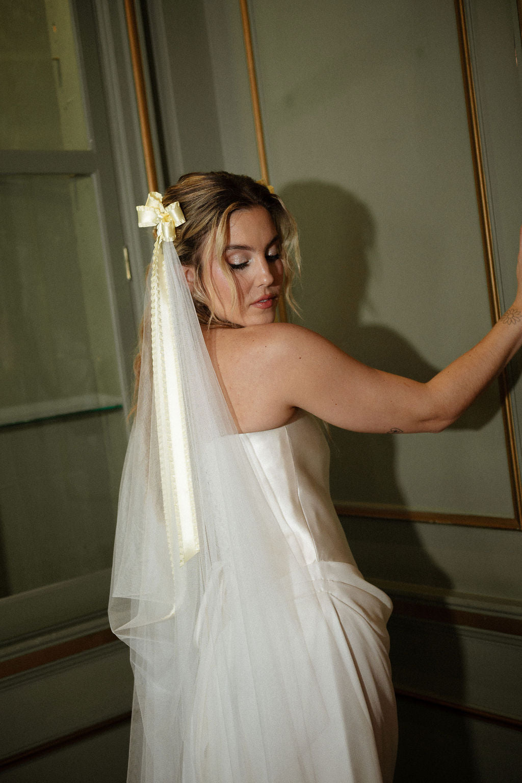 A woman wearing a strapless white wedding dress and a sheer veil with a cream bow, posing indoors against a paneled wall.