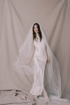 Woman wearing a white wedding dress with a long veil against a plain background