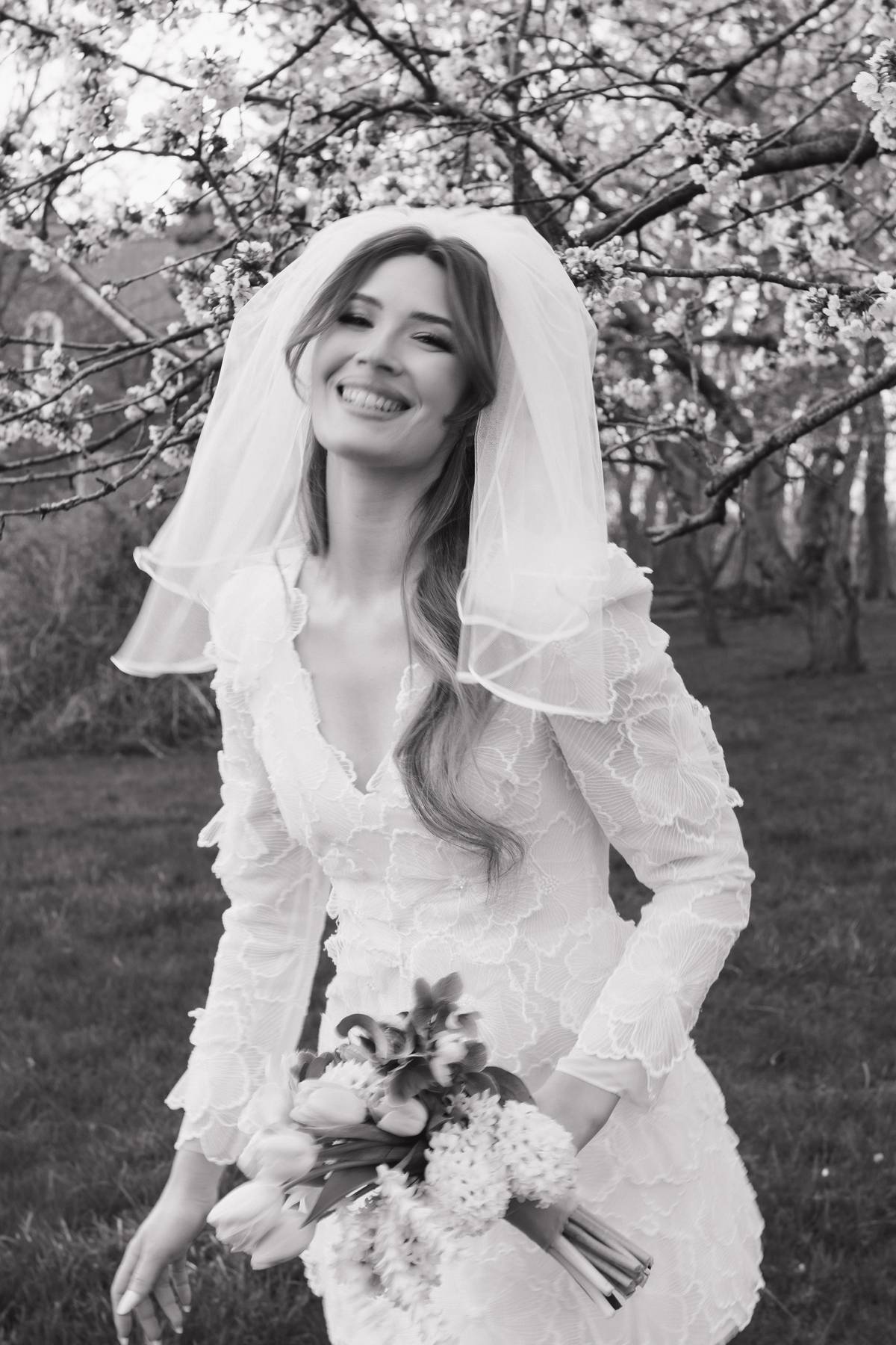 A black and white photograph of a smiling bride wearing a lace wedding dress and veil, holding a bouquet of flowers outdoors among blooming trees.