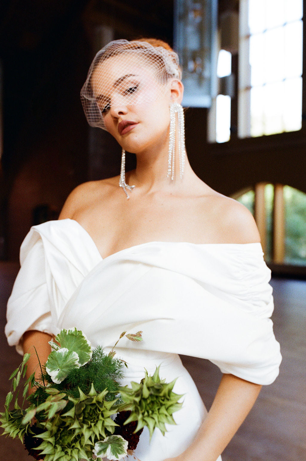 Bride in a white off-shoulder gown with a veil and green bouquet indoors.