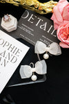 White bow earrings with pearls on a black card with floral and jewelry elements.