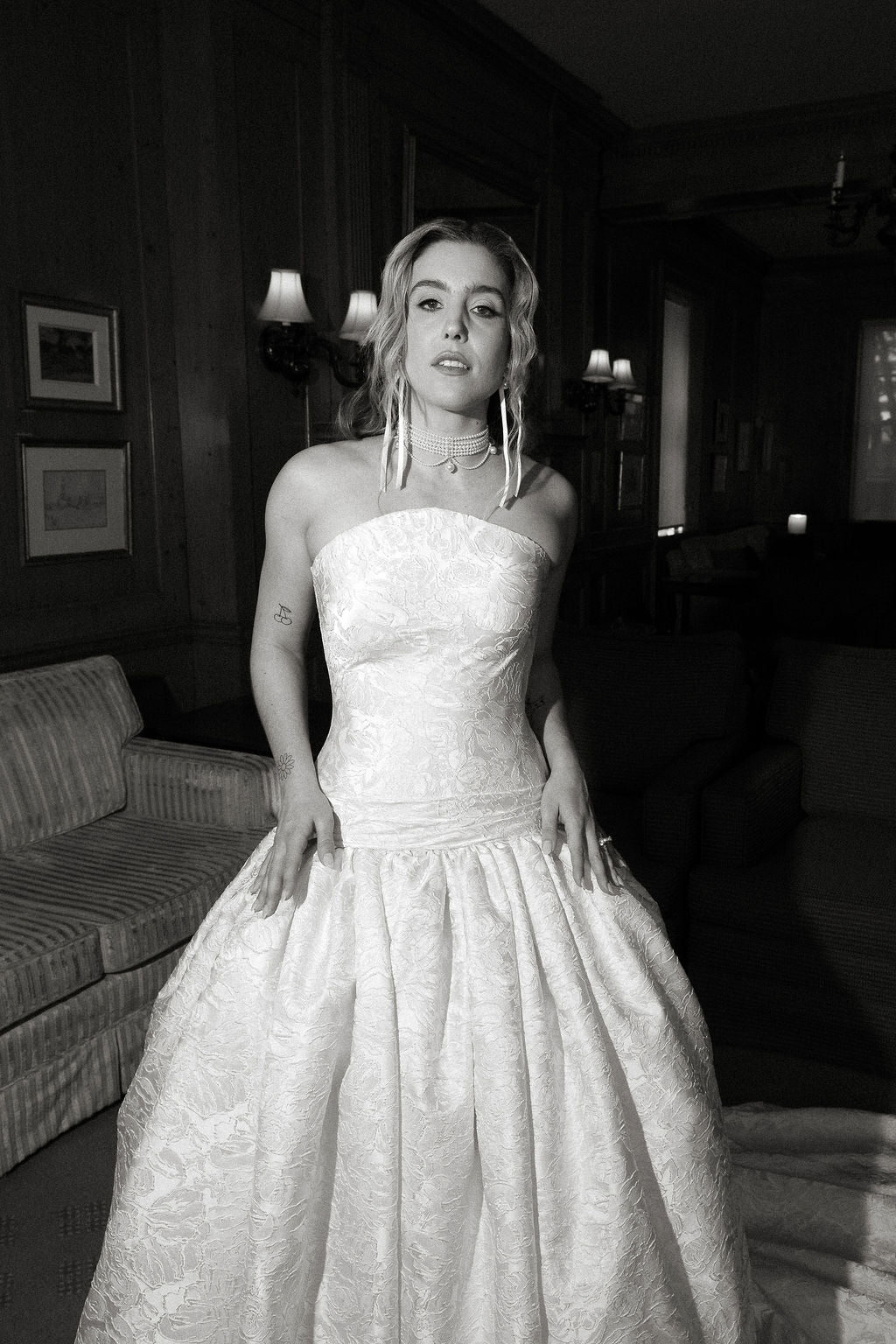 Woman in a strapless gown standing in an elegant room with framed pictures on the wall.