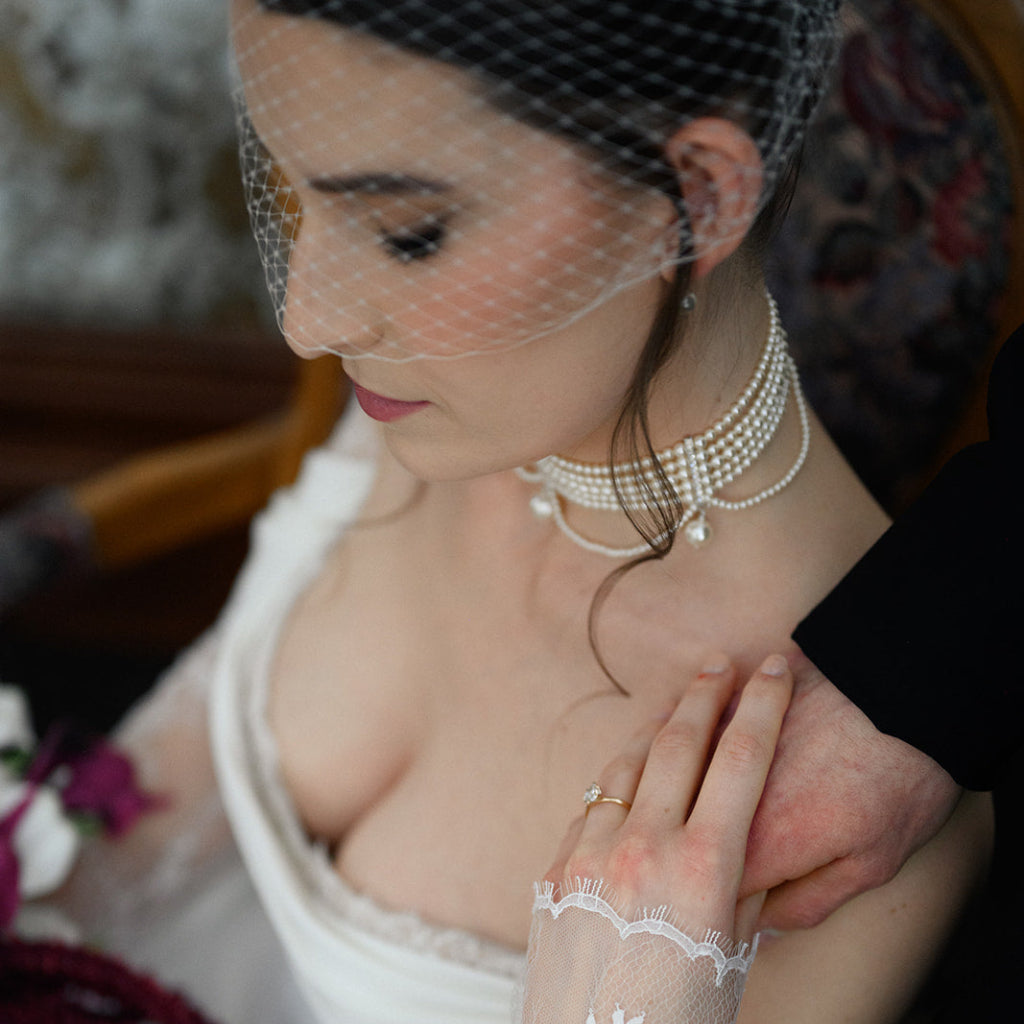 Woman in a wedding dress with lace gloves and pearl necklace, looking down.