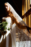 Woman in a white wedding dress with a lace veil, holding flowers, in front of a window.