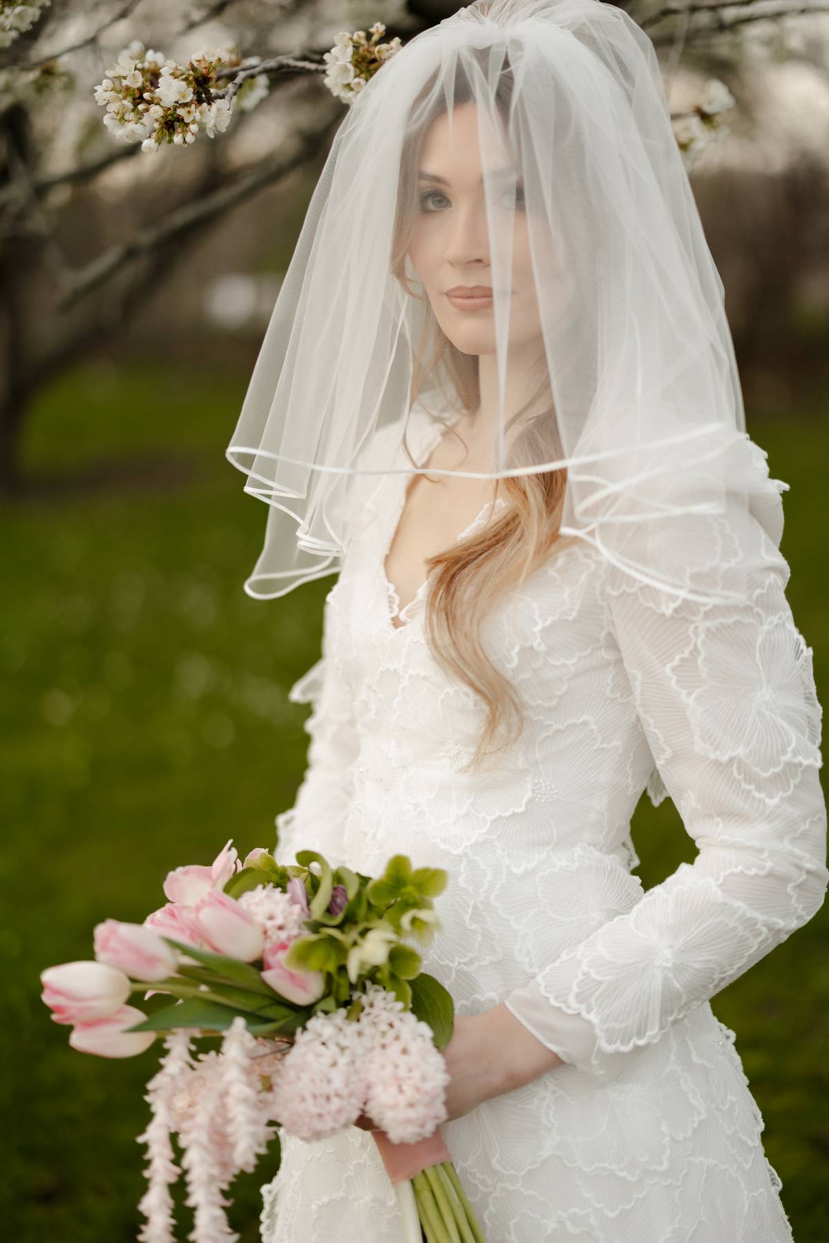 A woman wearing a white lace wedding dress and sheer veil holds a bouquet of pink tulips and pale pink flowers outdoors in a garden setting.
