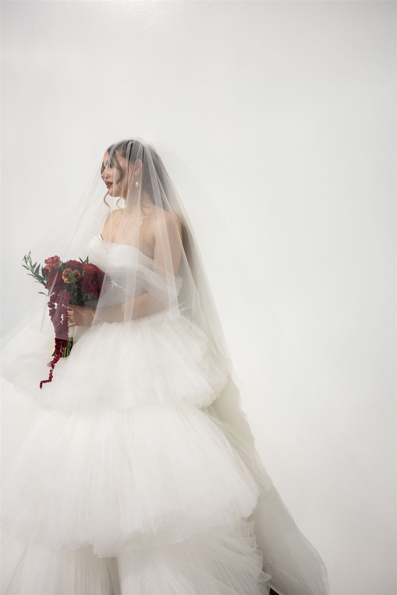 Bride in a white wedding dress with a veil holding a bouquet against a plain background