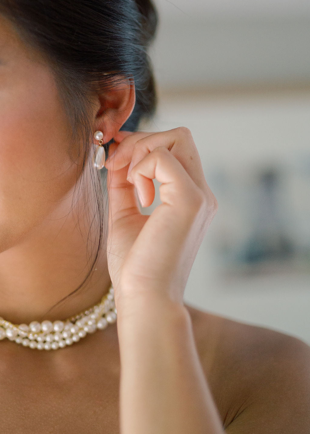 A close-up lifestyle shot of a woman wearing a delicate pearl drop earring and a multi-strand pearl necklace, adjusting her earring with her hand.