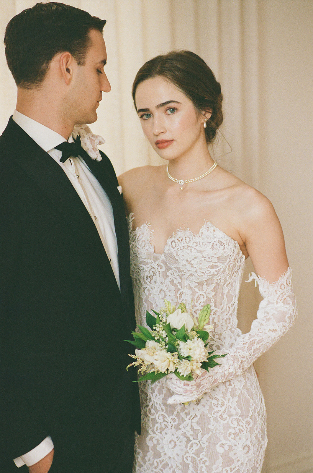 Man in tuxedo and woman in lace wedding dress with bouquet, standing close together.