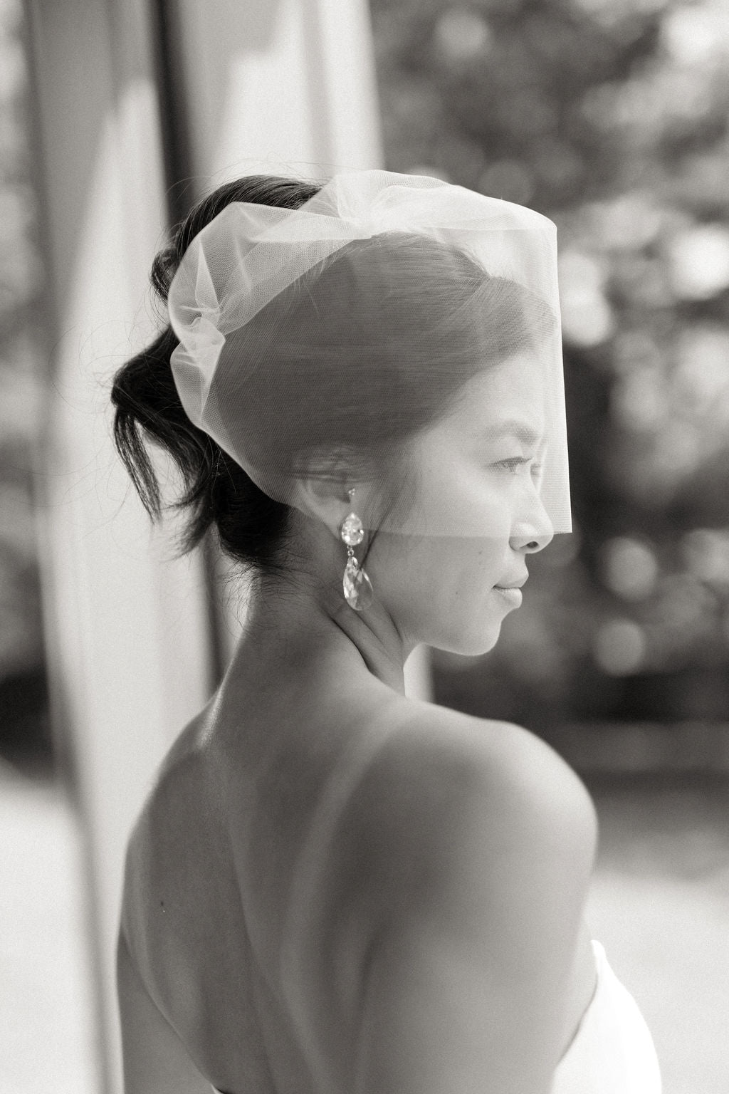 Black and white portrait of a woman in profile wearing a sheer white bridal veil and dangling earrings, with a softly blurred outdoor background.
