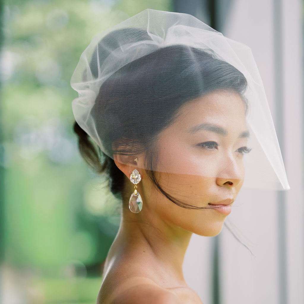A woman in profile wearing a sheer white bridal veil and white strapless dress, with dangling clear teardrop earrings, photographed against a softly blurred outdoor background.