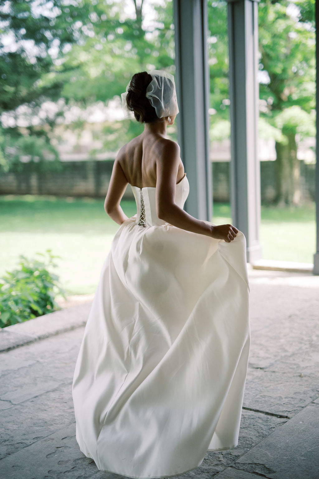 A bride in a strapless ivory wedding gown with a lace-up corset back and a sheer veil, photographed from behind while holding out the skirt on a stone patio with a garden background.