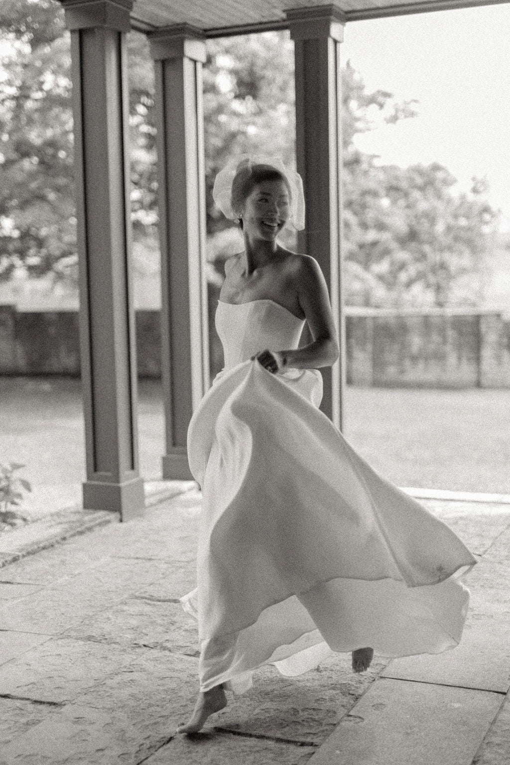 A woman in a flowing strapless white dress and matching headpiece dances barefoot on a stone patio under a covered porch with columns, captured in black and white.