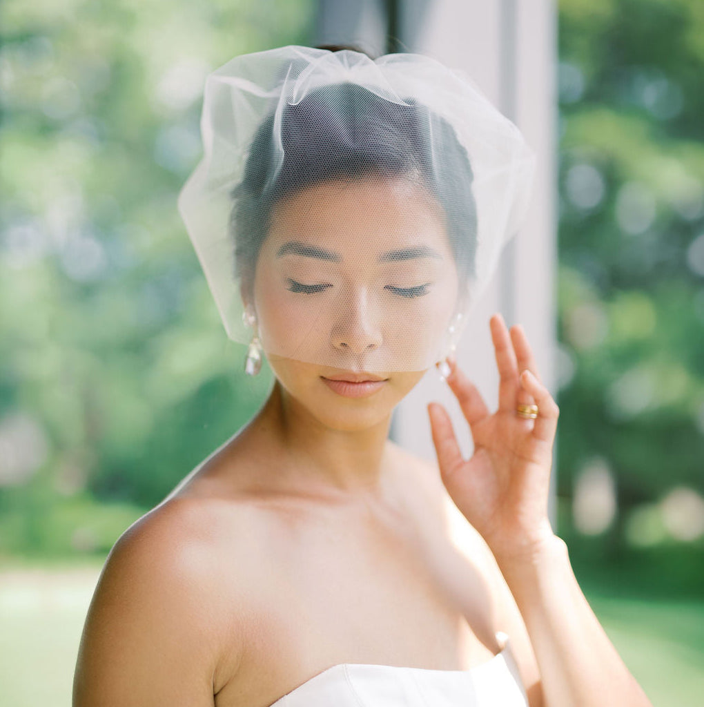 A woman wearing a white strapless dress and a sheer white veil, posed outdoors with a soft-focus green garden background.