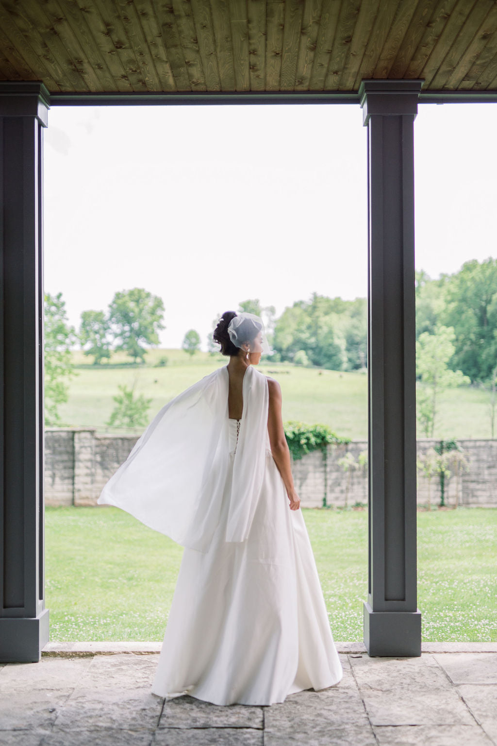 A bride in a sleeveless white wedding gown with a flowing veil stands with her back to the camera, framed by dark columns at an outdoor pavilion overlooking a green field.