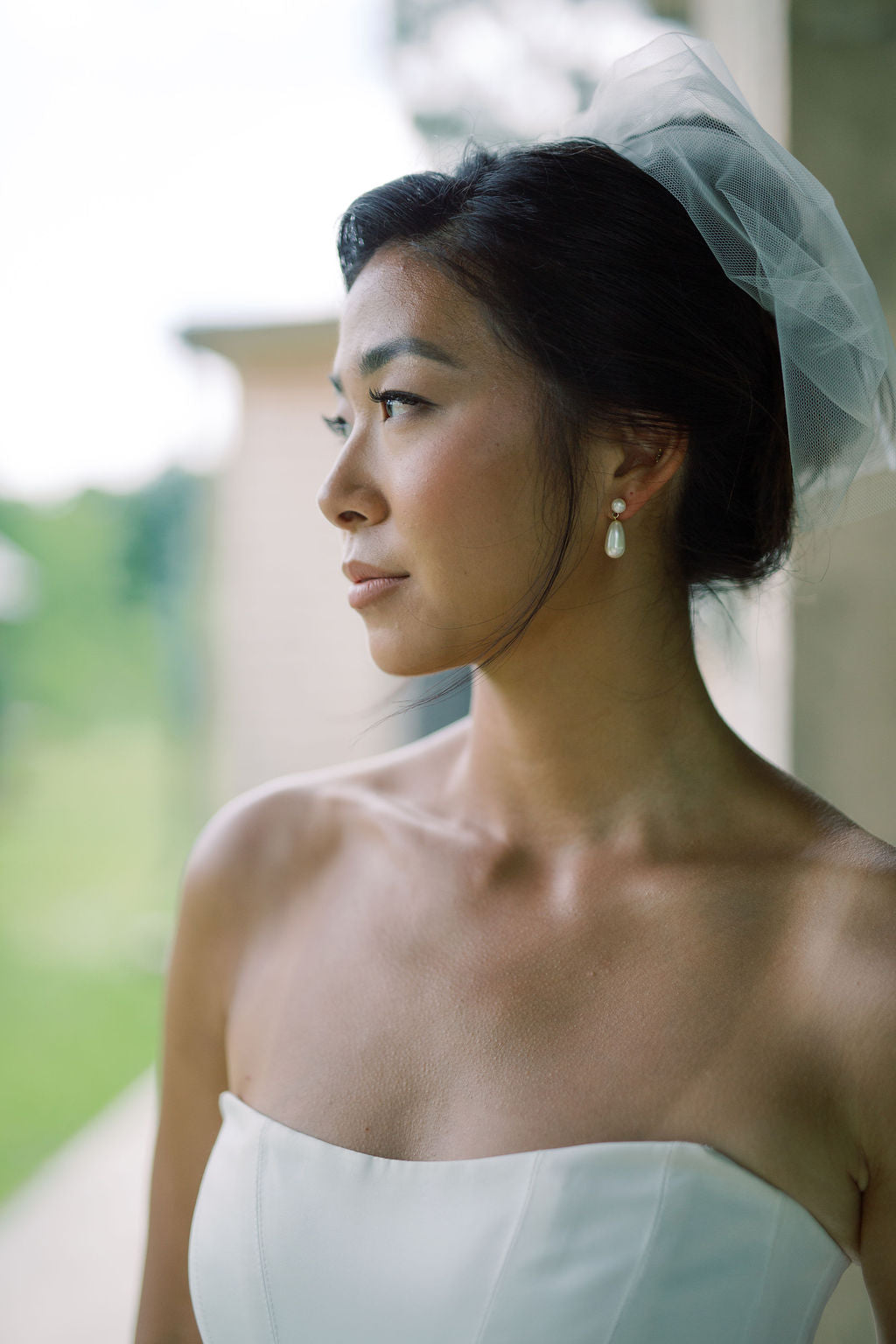 A woman in a strapless white wedding dress and sheer veil, shown in profile against a softly blurred outdoor background.