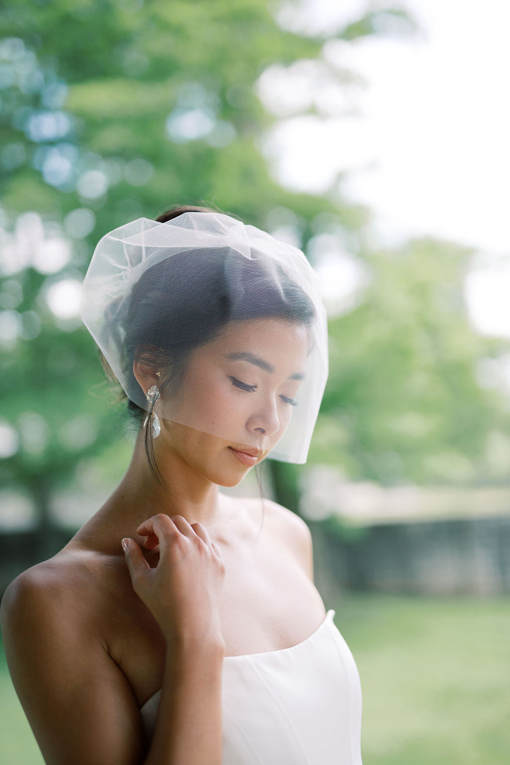 A woman outdoors wearing a white strapless dress and a sheer white bridal veil, with a softly blurred green garden background.