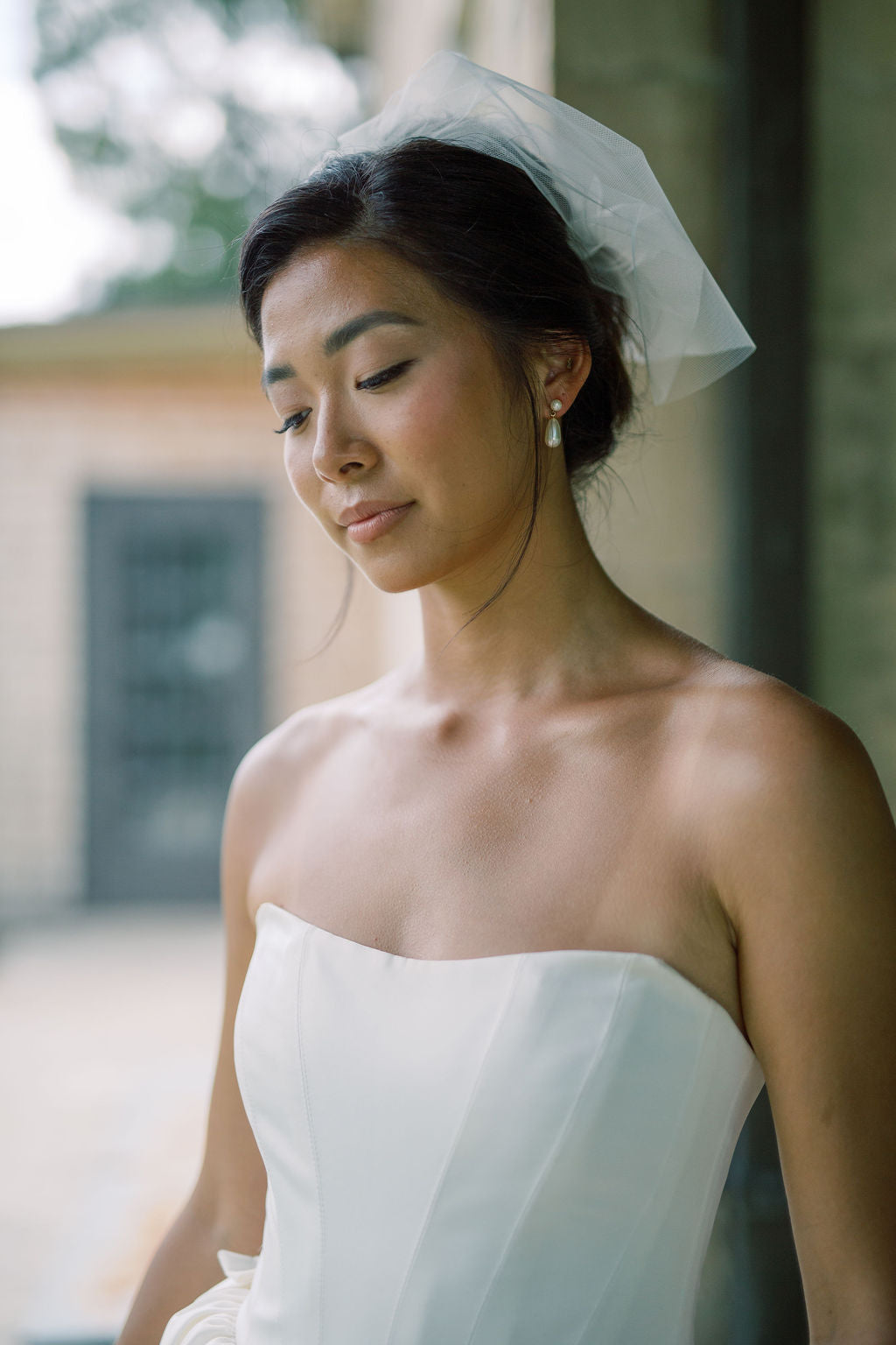 A woman wearing a strapless white wedding dress and a sheer white veil, photographed in a softly blurred outdoor setting.