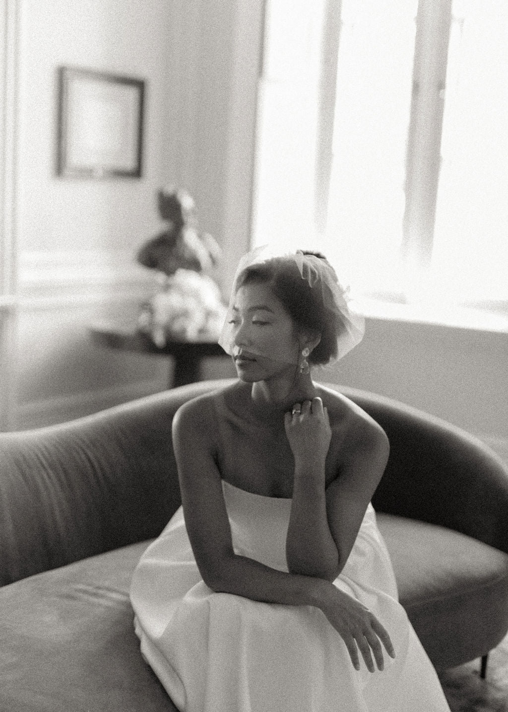 A black and white portrait of a woman in a strapless white dress sitting on a curved sofa in a softly lit room.